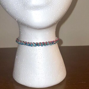 May include: A braided bracelet in shades of red, blue, and white is displayed on a white mannequin head. The bracelet is made of woven threads, showcasing a detailed pattern. The jewelry is a colorful accessory.