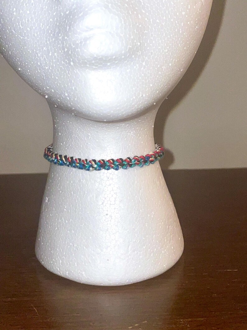 May include: A braided bracelet in shades of red, blue, and white is displayed on a white mannequin head. The bracelet is made of woven threads, showcasing a detailed pattern. The jewelry is a colorful accessory.