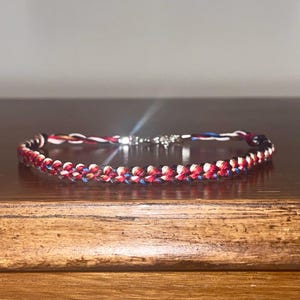 May include: A braided bracelet featuring red, white, and blue threads, with a silver clasp. The bracelet is displayed on a wooden surface, showcasing its woven design and color scheme. The bracelet's length is approximately 7 inches.