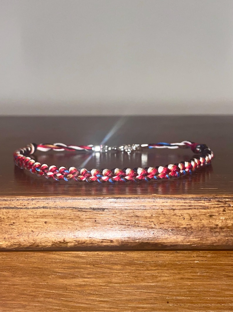 May include: A braided bracelet featuring red, white, and blue threads, with a silver clasp. The bracelet is displayed on a wooden surface, showcasing its woven design and color scheme. The bracelet's length is approximately 7 inches.