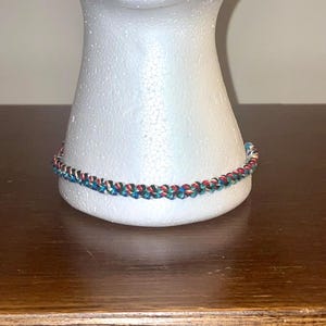 May include: A braided bracelet with red, green, blue, and white threads. The bracelet is displayed on a white mannequin head, resting on a wooden surface. The bracelet's design is a repeating pattern of the colors.