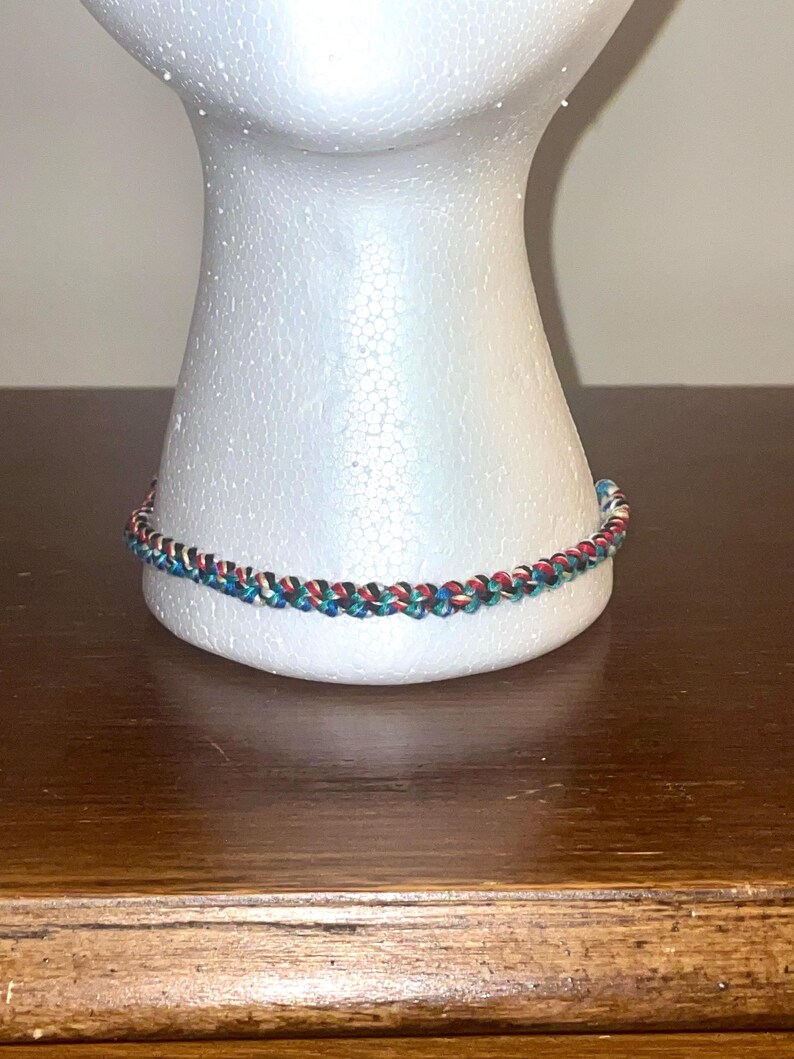 May include: A braided bracelet with red, green, blue, and white threads. The bracelet is displayed on a white mannequin head, resting on a wooden surface. The bracelet's design is a repeating pattern of the colors.