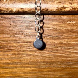 May include: Close-up of a silver-colored necklace chain with a small, round, gray pendant. The chain is made of small, interconnected links. The background is a wooden surface with horizontal grain.
