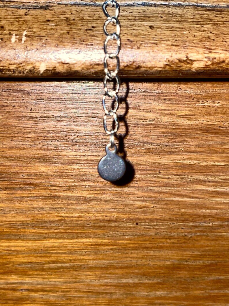 May include: Close-up of a silver-colored necklace chain with a small, round, gray pendant. The chain is made of small, interconnected links. The background is a wooden surface with horizontal grain.