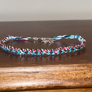 May include: Braided bracelet featuring a mix of red, black, blue, and white threads. The bracelet has a silver-colored clasp and a small decorative charm. The bracelet is displayed on a wooden surface.