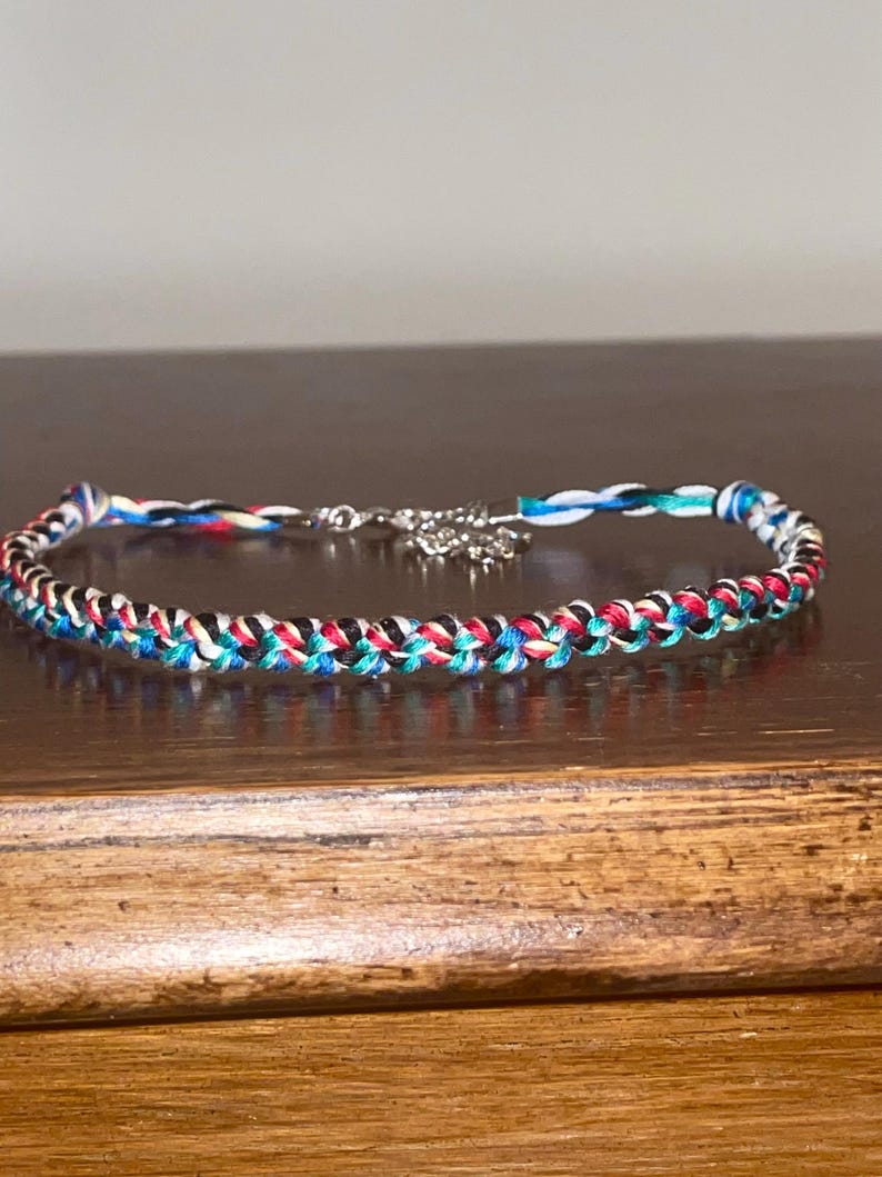 May include: Braided bracelet featuring a mix of red, black, blue, and white threads. The bracelet has a silver-colored clasp and a small decorative charm. The bracelet is displayed on a wooden surface.