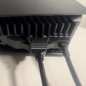 May include: A black electronic device featuring a series of vertical cooling fins along the top edge. Two black cables are plugged into the ports on the back panel, with one cable featuring a braided texture and a molded connector housing.