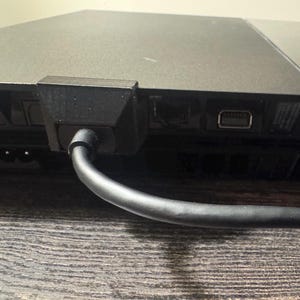 May include: A black plastic cable support bracket attached to the rear of a black gaming console. The bracket secures a black power cable that extends outward. The console features a rectangular port and a two-pin power input socket nearby.