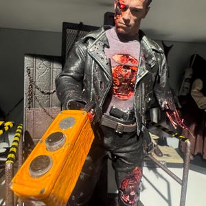 May include: A detailed action figure of a cyborg from a science fiction film. The figure wears a black leather jacket, black pants, and a gray shirt. The figure is holding an orange control box with two silver buttons. The figure has exposed mechanical parts.