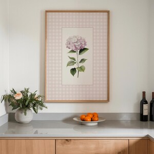 Op de afbeelding: Een ingelijste bloemenprint met een roze hortensia en groene bladeren, tegen een roze en wit geruit patroon. De lijst is van licht hout. Een vaas met bloemen en een schaal met sinaasappels staan op een aanrecht in een keuken.