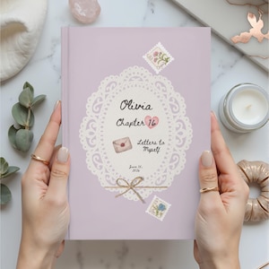 May include: A lavender journal titled "Olivia Chapter 16 Letters to Myself." The cover features a white lace design, a heart, and postage stamp illustrations. The journal is held open by hands with gold rings.