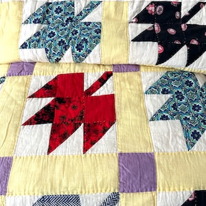 May include: A close-up of a handmade quilt with leaf-shaped patterns. The quilt features a patchwork design with red, blue, and black leaf motifs set against a yellow and white background. The quilt is a handmade textile.