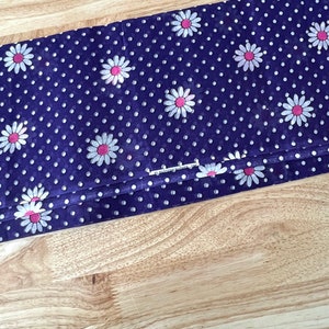 Peut inclure: Tissu pli&eacute; avec un fond bleu fonc&eacute;, recouvert de pois blancs et de motifs de fleurs de marguerite &agrave; centres roses. Le tissu est pli&eacute; sur lui-m&ecirc;me, montrant le motif en d&eacute;tail. Le tissu est sur une surface en bois brun clair.