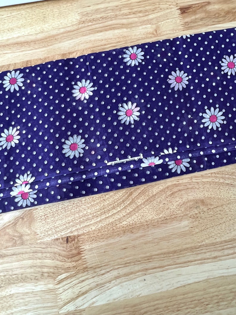 Peut inclure: Tissu pli&eacute; avec un fond bleu fonc&eacute;, recouvert de pois blancs et de motifs de fleurs de marguerite &agrave; centres roses. Le tissu est pli&eacute; sur lui-m&ecirc;me, montrant le motif en d&eacute;tail. Le tissu est sur une surface en bois brun clair.