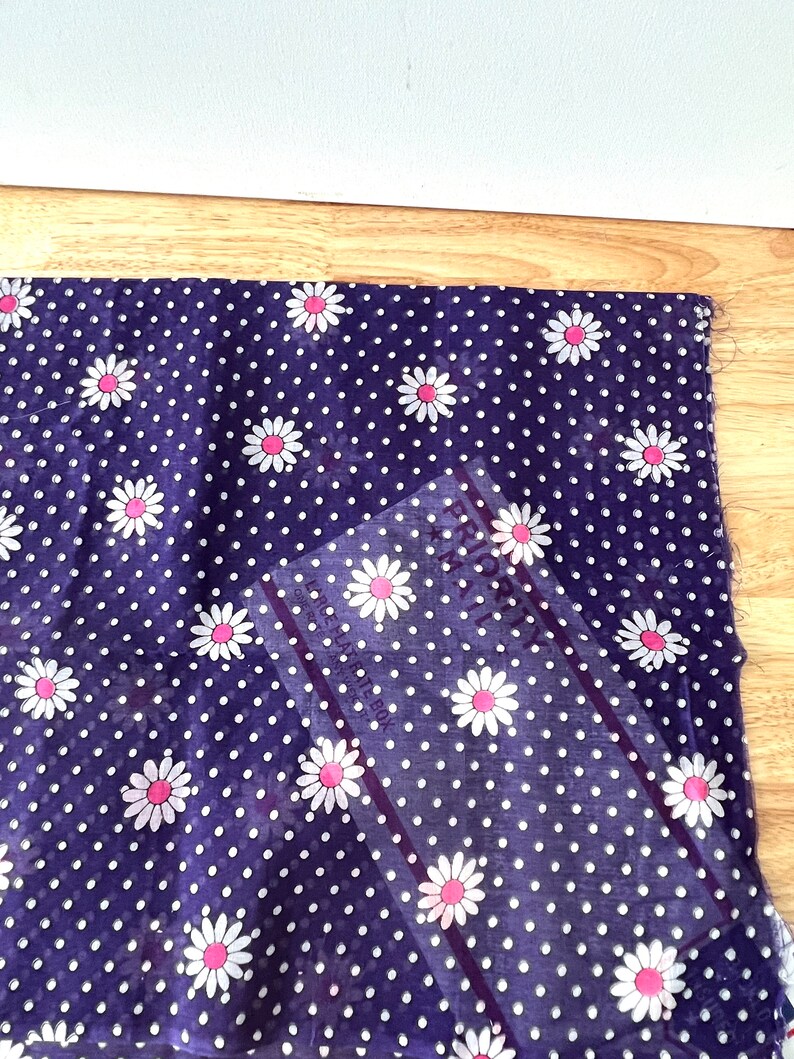Peut inclure: Un morceau de tissu bleu marine avec un motif de pois blancs et des marguerites blanches &agrave; centres roses. Une r&egrave;gle transparente est plac&eacute;e en diagonale sur le tissu. Le tissu est sur une surface en bois brun clair.
