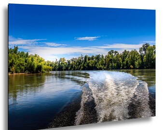 May include: A scenic landscape photograph of a river with a wake from a boat. The water is dark blue, reflecting the bright blue sky with scattered clouds. Lush green trees line the riverbanks, creating a serene and natural environment.