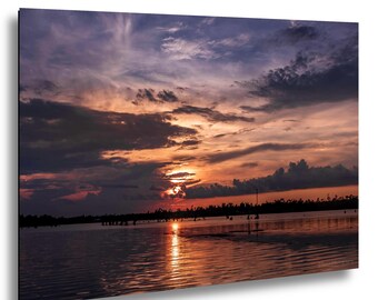 May include: A framed print of a sunset over a body of water. The sky is filled with shades of purple, orange, and gray clouds. The sun is setting on the horizon, reflecting on the water's surface. The print is mounted on a black frame.