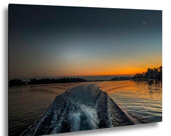 May include: A scenic canvas print of a sunset over a river. The wake of a boat cuts through the water, reflecting the orange and yellow hues of the sky. A crescent moon is visible in the upper right corner, with dark trees lining the horizon.