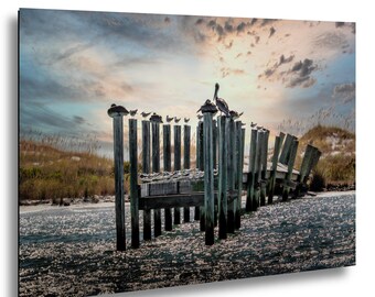 May include: A rectangular wall art print featuring a row of weathered wooden pilings extending into dark water. Numerous birds are perched atop the posts under a soft, cloudy sky at sunset, with sand dunes visible in the background.