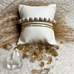 Peut inclure: Trois bracelets avec des perles blanches et dorées sont présentés sur un coussin blanc. Deux sont des chaînes dorées, et l'un comporte des perles de pierre blanches avec des accents dorés. Des cristaux clairs et ambrés entourent le coussin.