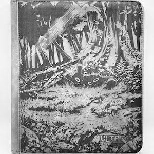May include: A gray, leather-bound journal featuring a detailed black and white illustration of a forest scene with stylized creatures. The cover art depicts a forest with trees, ferns, and a ray of light. The journal has a smooth, gray border.