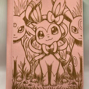 May include: Pink card binder with a gold illustration of three cartoon creatures in a grassy field. The central creature has a bow and ribbons. The illustration is detailed with fine lines and shading.