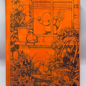 May include: Orange leather journal with an engraved illustration. The design features a cartoon character in a window with flames, birds, flowers, and a potted plant. The journal has a vertical orientation and a textured surface.