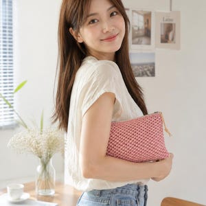 May include: A pink woven clutch bag with a zip and leather pull. The bag is held by a person wearing a white top and blue jeans. The background includes a vase of flowers and a magazine.