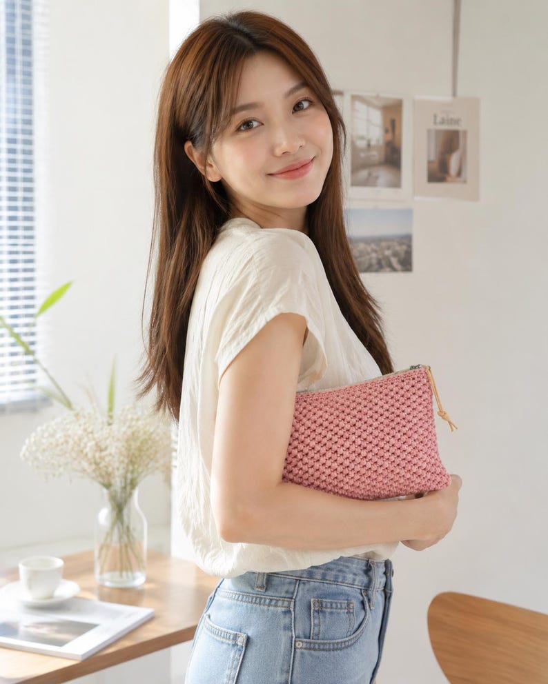 May include: A pink woven clutch bag with a zip and leather pull. The bag is held by a person wearing a white top and blue jeans. The background includes a vase of flowers and a magazine.