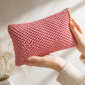 May include: A handmade, pink crochet clutch with a textured stitch pattern. The rectangular bag has a zip closure with a tan pull tab. The clutch is held in front of a white background.