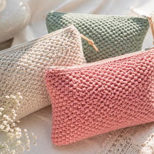 May include: Three crochet pouches in cream, sage green, and pink. Each pouch has a textured stitch, a zip closure, and a small leather pull. The rectangular pouches are made from cotton yarn. The image shows a close-up of the pouches.