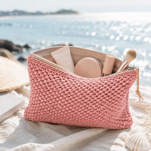 May include: A pink, textured cosmetic bag with a zip, containing makeup items. A straw hat and a book are beside the bag. The background shows the sea and a sunny day.