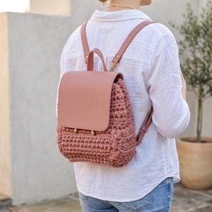 May include: A dusty rose backpack featuring a smooth leather-like flap and a textured, chunky crocheted body. The bag includes matching adjustable shoulder straps, a top handle, and gold-toned metal hardware accents on the front closure.