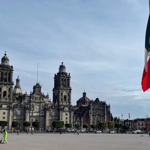 Puede incluir: Vista de la Catedral Metropolitana de la Ciudad de México, un gran edificio de piedra con múltiples torres y cúpulas. La bandera mexicana ondea en primer plano. Gente caminando en la plaza.