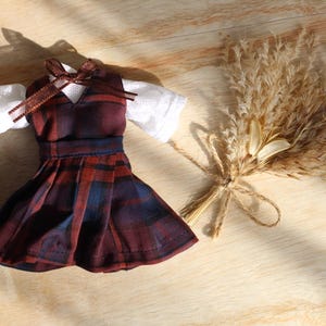 May include: A miniature dress in a burgundy, navy, and red plaid pattern, with a white blouse. A brown ribbon adorns the neckline. A bundle of dried wheat tied with twine is next to the dress, on a light wooden surface.