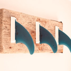 Peut inclure: Un support mural pour ailerons de planche de surf, composé d'une planche de bois vieillie. Trois ailerons bleu-vert sont fixés par des supports blancs. Le bois présente un aspect naturel et rustique, avec des veines et des nœuds visibles. Un objet fonctionnel et décoratif.