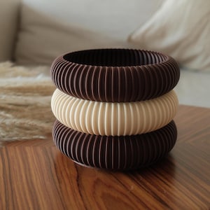 Könnte beinhalten: Drei gestapelte, gerippte Armreifen in Braun- und Cremetönen. Die oberen und unteren Armreifen sind dunkelbraun, während der mittlere cremefarben ist. Die Armbänder bestehen aus einem festen Material und haben eine geriffelte Textur.