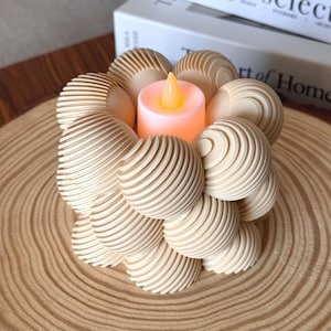 May include: A decorative wooden candle holder with a light pink LED candle. The holder is made of multiple wooden spheres with a grooved design. The candle holder sits on a wooden slice.