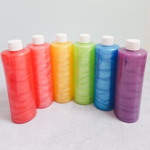 May include: Six bottles of iridescent liquid in a rainbow of colors: red, orange, yellow, green, blue, and purple. The bottles are lined up in a row on a white surface.