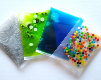 May include: Four clear plastic bags filled with different sensory materials. One bag contains silver glitter, another has green goo with colorful eyes, a third has blue liquid, and the last bag has colorful beads.
