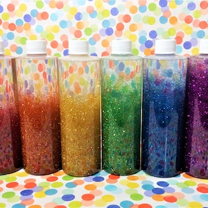 May include: Ten clear plastic bottles filled with colorful glitter and water. The bottles are lined up in a row on a white background with colorful polka dots.