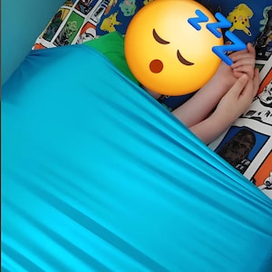 May include: A bright turquoise fitted sheet covering a bed with a patterned pillowcase featuring various cartoon characters. The sheet is pulled up over the bed surface, creating a smooth, solid-colored fabric layer across the mattress area.