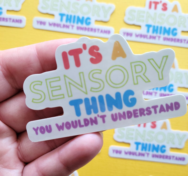 May include: A white sticker with the text "It's a Sensory Thing You Wouldn't Understand" in rainbow colors. The sticker is held in a hand.