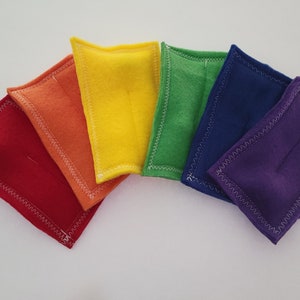 May include: Six small, rectangular fabric pouches in a rainbow color scheme. The pouches are sewn with a white zig-zag stitch around the edges. The pouches are arranged in a semi-circle, with the red pouch at the bottom and the purple pouch at the top.