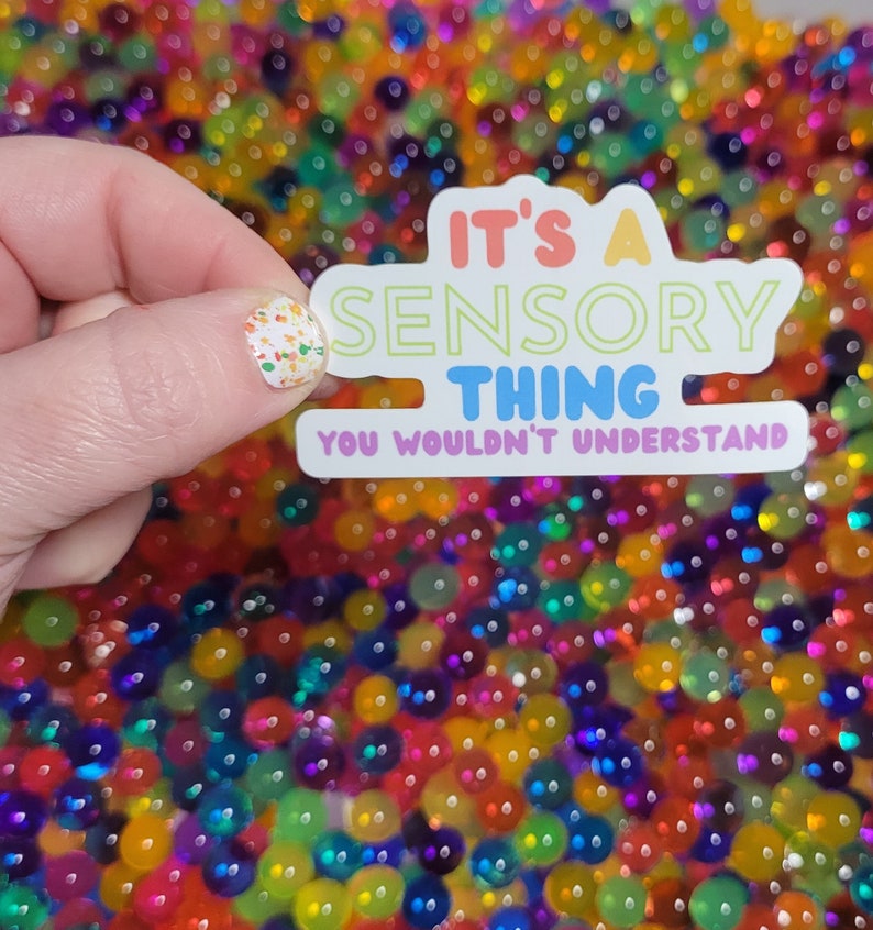 May include: White sticker with the text "It's a Sensory Thing You Wouldn't Understand" in rainbow colors. The sticker is held by a hand against a background of colorful iridescent beads.