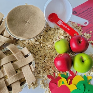 May include: An overhead shot displays a sensory play kit with felt pie crusts, red and green apples, oats, a measuring spoon, and a recipe card. The kit includes a white bowl and a red measuring spoon with "Betty Crocker" printed on it.