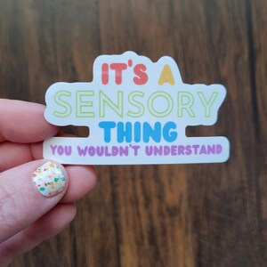 May include: A white sticker with the text "It's a Sensory Thing You Wouldn't Understand" in rainbow colors. The sticker is held by a hand with colorful nail polish.