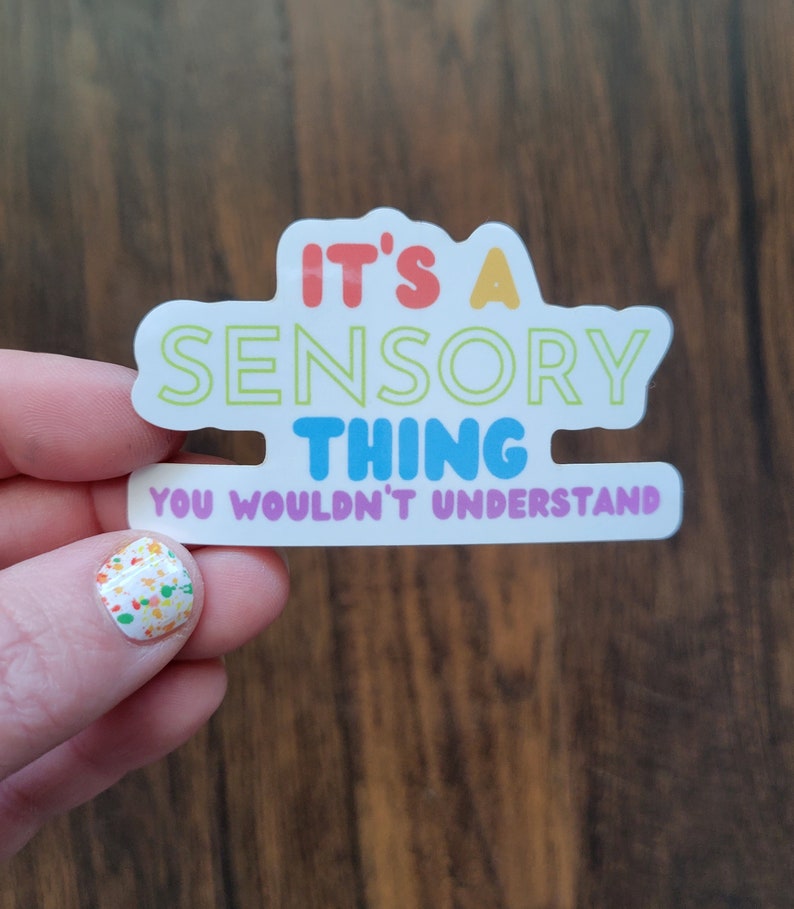 May include: A white sticker with the text "It's a Sensory Thing You Wouldn't Understand" in rainbow colors. The sticker is held by a hand with colorful nail polish.