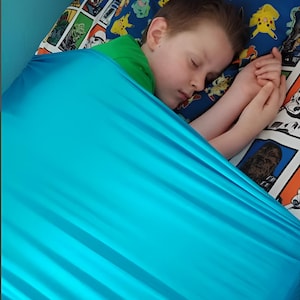 May include: A child sleeping in a turquoise weighted blanket on a bed with a colorful cartoon patterned bedding.