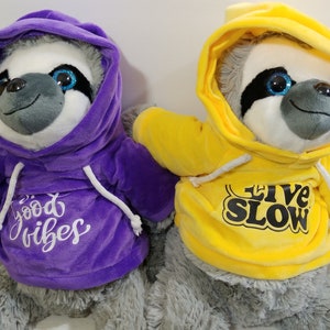 May include: Two plush sloth toys wearing hoodies. One sloth has a purple hoodie with "good vibes" text, and the other has a yellow hoodie with "live slow" text. Both sloths have gray fur and large, sparkly blue eyes.